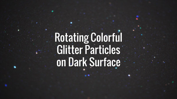 Seamlessly looping spinning flickering multicolored glitter particles on dark surface.